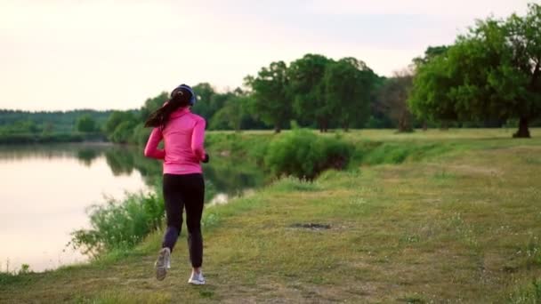 Brunetka z długimi włosami w słuchawkach biegnie wzdłuż rzeki w parku rano o świcie, w lecie w różowy kurtkę i czarne spodnie - Materiał filmowy, wideo