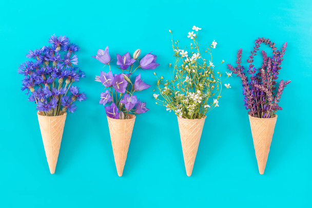 vier Waffelzapfen mit Thymian, Kornblume, blauen Glocken und weißen Blütensträußen auf blauer Oberfläche. flache Lage, von oben betrachtet floraler Hintergrund. - Foto, Bild