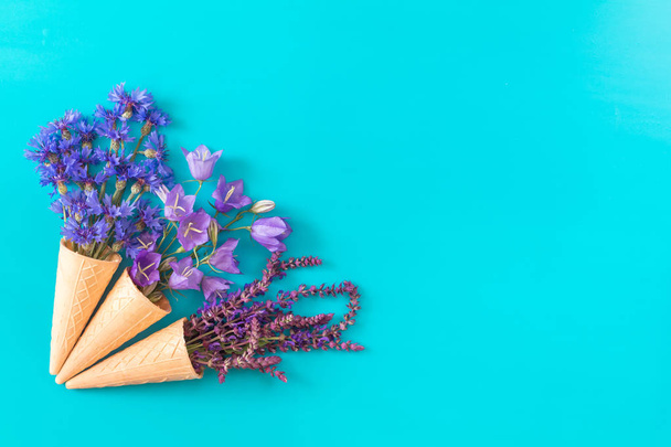 drei Waffelzapfen mit Thymian, Kornblume, blauen Glocken und weißen Blütensträußen auf blauer Oberfläche. flache Lage, von oben betrachtet floraler Hintergrund. - Foto, Bild