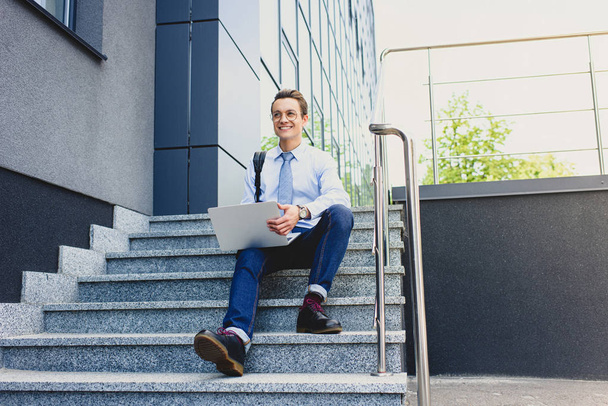 guapo sonriente joven freelancer en gafas sentado en las escaleras y el uso de la computadora portátil
   - Foto, Imagen