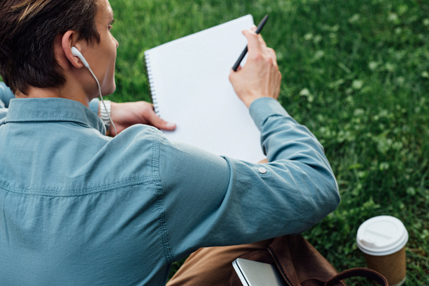 cropped shot of man in earphones writing in blank notebook while sitting on grass - Photo, Image