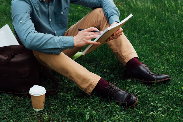 cropped shot of young man writing in notebook while sitting on grass - Foto, Imagem