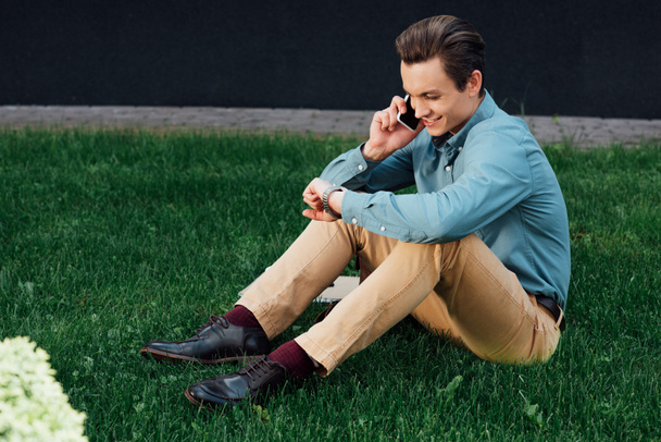 Smartphone ile konuşmak ve çim üzerinde otururken kol saati kontrol genç adam gülümseyen - Fotoğraf, Görsel