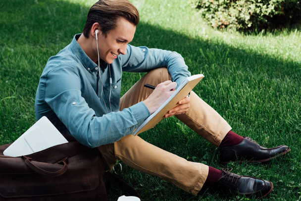 high angle view of smiling young man in earphones taking notes in notepad - Photo, Image