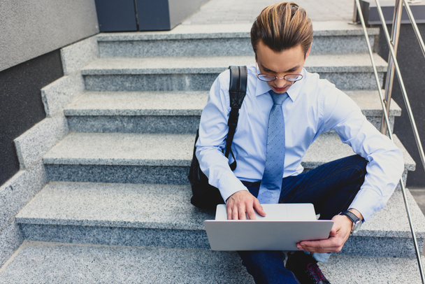 красивый молодой бизнесмен в очках сидит на лестнице и пользуется ноутбуком
 - Фото, изображение