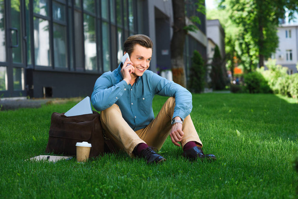 ハンサムな若い男性のフリーランサーの芝生の上に座っているとスマート フォンで話を笑って - 写真・画像