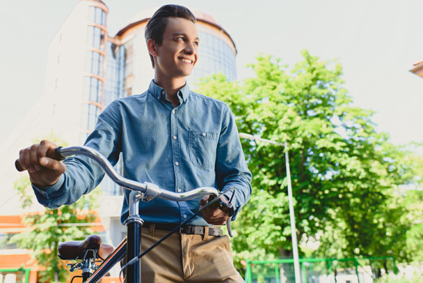 Tiefansicht eines gut aussehenden, lächelnden jungen Mannes, der mit Fahrrad auf der Straße steht und wegschaut - Foto, Bild