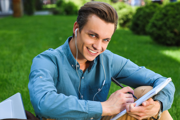 pohledný mladý muž v sluchátka zapisování poznámek a usmívá se na kameru - Fotografie, Obrázek