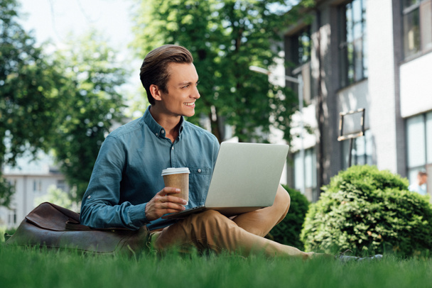 usmíval se mladý muž, držící pohárek a pomocí přenosného počítače, zatímco sedí na trávě - Fotografie, Obrázek