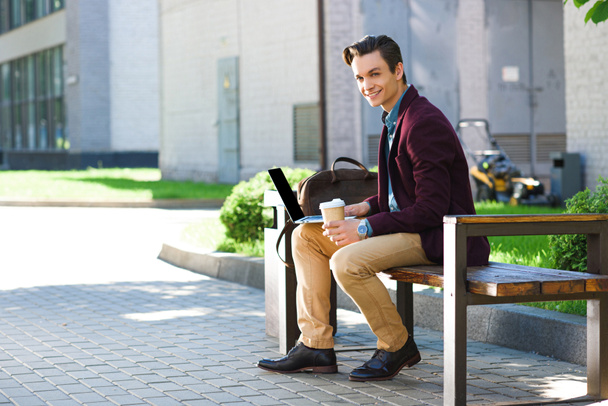 handsome young freelancer using laptop and smiling at camera while sitting on bench - Valokuva, kuva