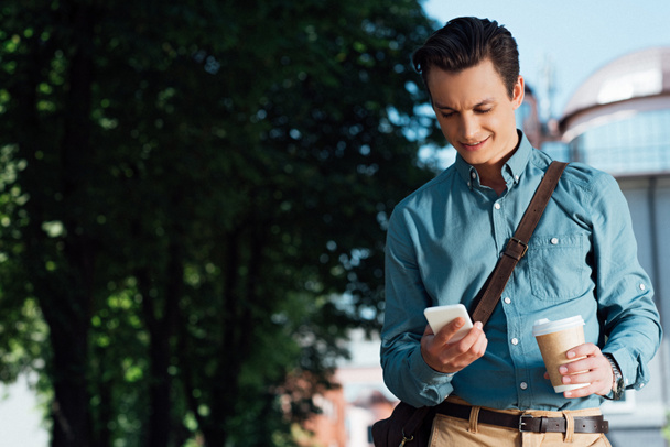 komea hymyilevä nuori mies pitää paperimukia ja käyttää älypuhelinta
 - Valokuva, kuva