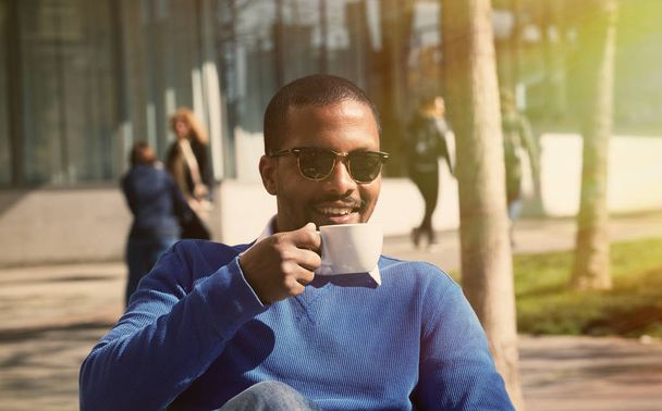 Portrait of smiling confident young African-American businessman in formal wear sitting at bench in city park and drinking hot coffee.Blurred background.Horizontal. - Φωτογραφία, εικόνα