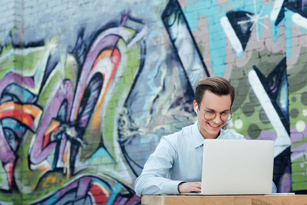 sonriente joven freelancer en gafas usando laptop mientras está sentado afuera
 - Foto, Imagen