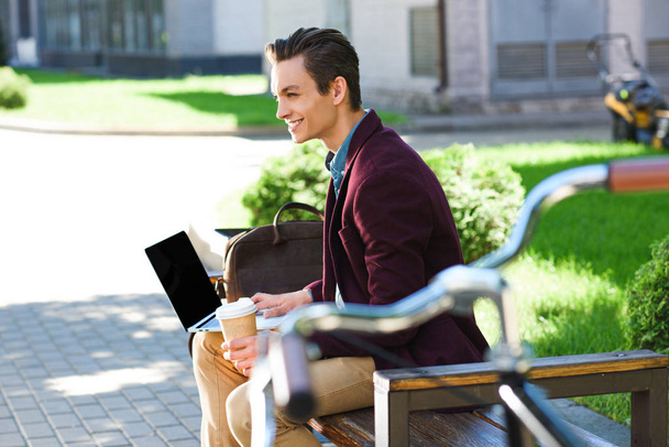 笑顔の若い男がベンチに空白の画面を持つノート パソコンを使用しての側面図 - 写真・画像