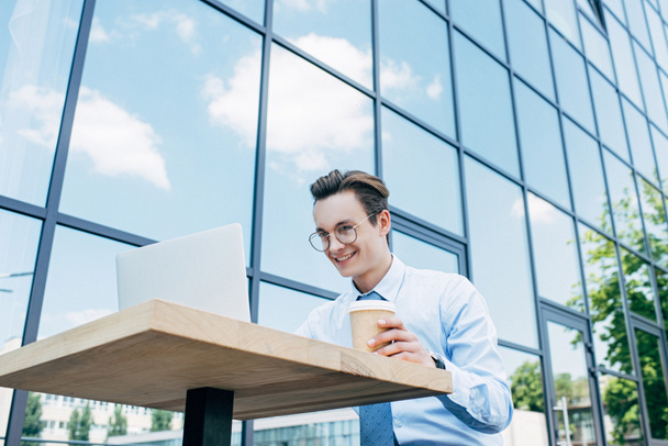 low angle view of smiling young freelancer holding paper cup and using laptop outside modern building - Photo, Image