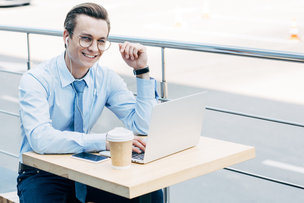 smiling young businessman in eyeglasses using laptop and earphones outside - Photo, Image