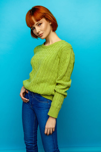 Colorful portrait of young female model with red hair and green sweater isolated on blue background - Foto, Imagem