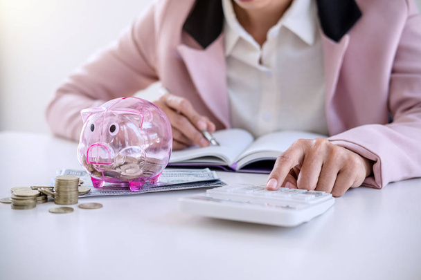 Business woman putting coins into piggy bank, writing to report and using calculator to analysis business investment strategy with income of money coin and dollar, financial concept. - Fotó, kép