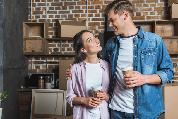 joven pareja interracial con tazas de papel de café de pie en la cocina mientras se muda a un nuevo hogar y se mira entre sí
 - Foto, Imagen