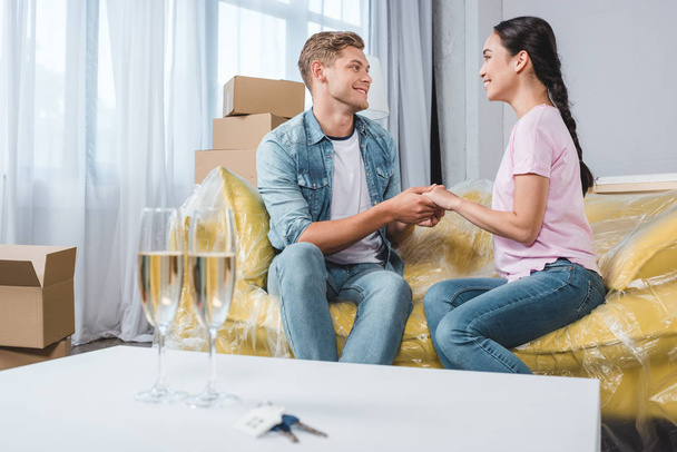 beau jeune couple assis sur le canapé et tenant la main après avoir emménagé dans une nouvelle maison avec des verres de champagne au premier plan
 - Photo, image