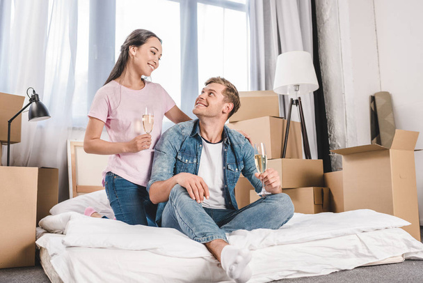 beautiful young couple sitting on bed with champagne while moving into new home - Photo, Image