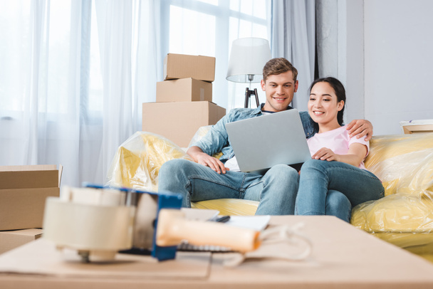 beautiful young couple using laptop on couch while moving into new home - Photo, Image