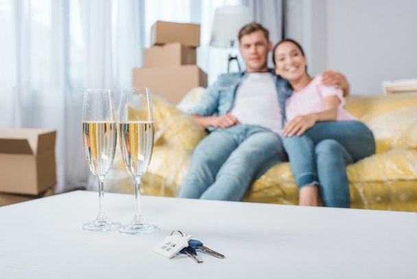 hermosa pareja joven relajarse en el sofá mientras se muda a un nuevo hogar con copas de champán y llaves en primer plano
 - Foto, Imagen