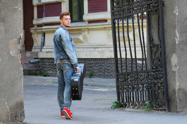Молодой веселый парень с рыжими волосами и бородой у ворот с гитарой в футляре
 - Фото, изображение