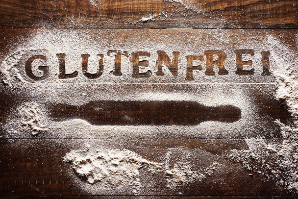 Aufnahme eines mit glutenfreiem Mehl bestreuten Holztisches, auf dem neben der Silhouette eines Nudelholzes der Text glutenfrei zu lesen ist - Foto, Bild