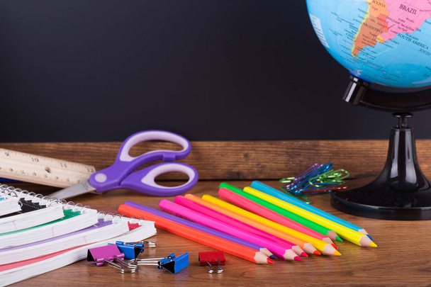 Assortment of back to school supplies on a wooden desktop with a blank chalkboard in background - Фото, изображение