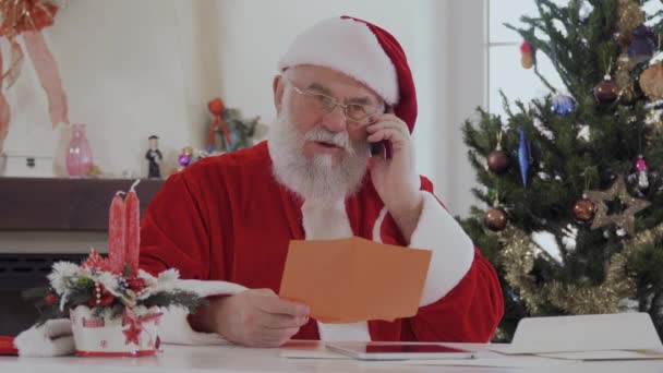 Santa Claus with serious face in red costume near bright decorated fir tree and fireplace talking on the cell phone. Old man is serious and focused on conversation. On the table there are two red - Video
