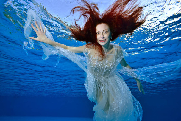 A girl with red hair in a white dress swims and plays underwater on a blue background, looks at the camera and smiles. Portrait. Bottom view from under the water. Horizontal orientation. - 写真・画像