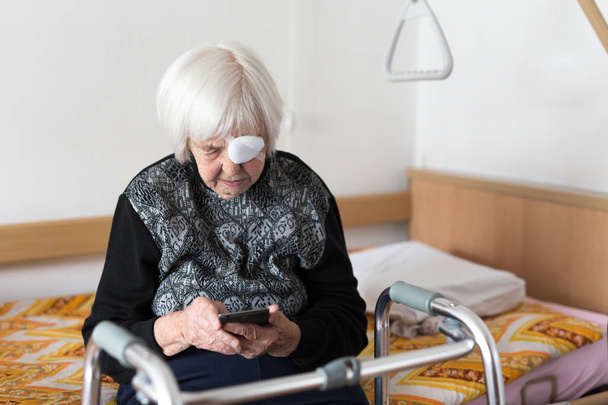Μόνος ηλικιωμένους 95 χρόνια γριά γυναίκα κάθεται στο το κακό χρησιμοποιώντας μοντέρνο κινητό τηλέφωνο. - Φωτογραφία, εικόνα