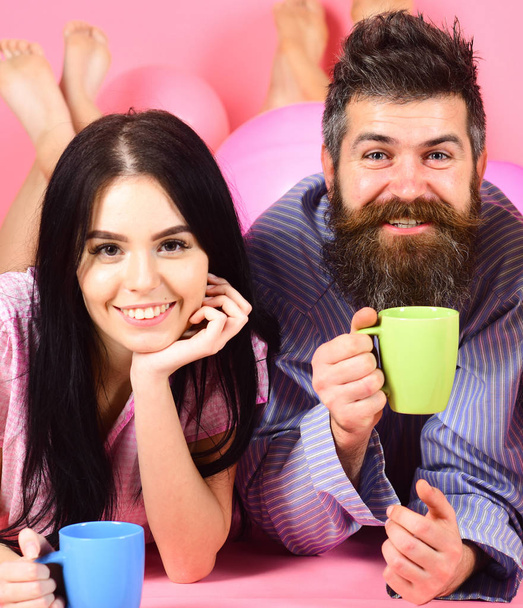 Man and woman on smiling faces lay, pink background. Couple in love drink coffee in bed. Man and woman in domestic clothes, pajamas. Couple relax in morning with coffee. Family tradition concept - Photo, image