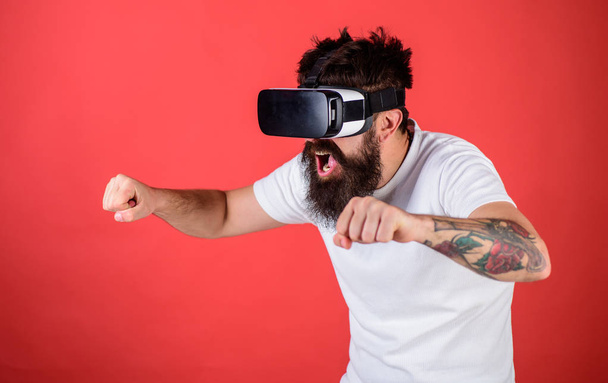 Hipster on excited face driving bike on high speed in virtual reality with modern digital gadget. Man with beard in VR glasses driving motorbike, red background. Virtual driving lessons concept - Photo, Image