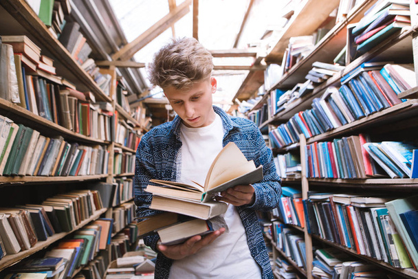 Beau jeune homme se tient dans une vieille bibliothèque publique avec des livres dans ses mains et lit. Un étudiant est à la recherche de littérature à la bibliothèque. Concept d'auto-apprentissage
. - Photo, image