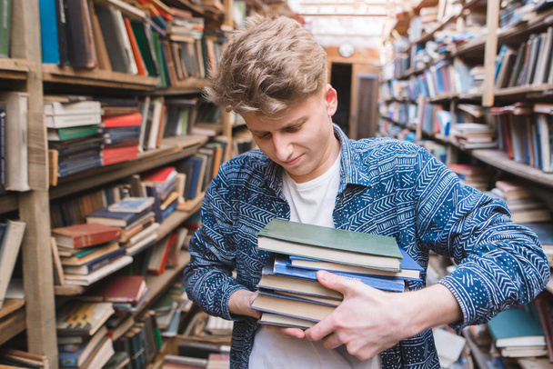 Young student with a lot of books in the hands of the library. A man is hard at the books in his hands. Portrait of a tired student with heavy books in his hands. - Foto, Imagen