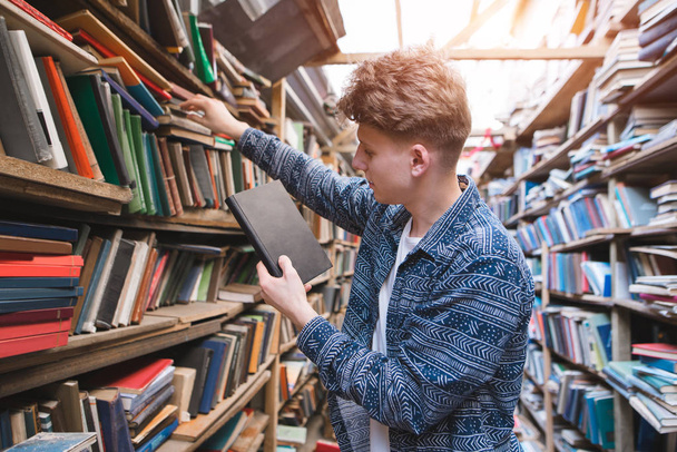 Knappe jongeman op zoek naar boeken over boekenkasten van een oude bibliotheek. Portret van een student die boeken uit de schappen van de openbare bibliotheek bladert. - Foto, afbeelding