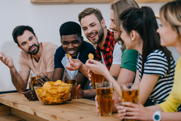 smiling group of multicultural friends eating chips, drinking beer and watching soccer match at bar  - Фото, изображение