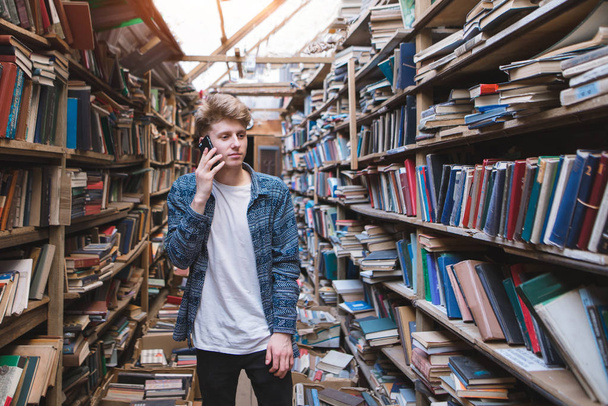 Μαθητής στέκεται στη βιβλιοθήκη κοντά σε ράφια και μιλάει μέσω τηλεφώνου. Τηλεφωνικές συνομιλίες στη βιβλιοθήκη. - Φωτογραφία, εικόνα