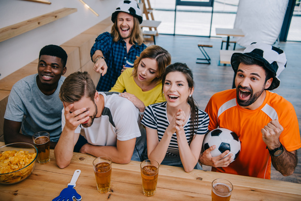 vista de ángulo alto de amigos multiculturales sonrientes en sombreros de pelota de fútbol sentado en el mostrador del bar con la bola, vasos de cerveza, tazón con patatas fritas y aplausos de mano durante el reloj del partido de fútbol
  - Foto, imagen