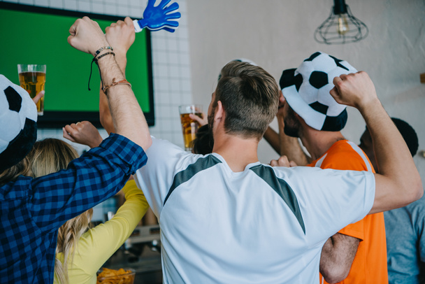rear view of football fans in soccer ball hats celebrating with hand clappers and doing yes gestures during watch of soccer match on tv screen at bar  - Photo, Image
