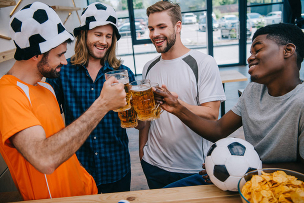 smiling multicultural group of male football fans clinking beer glasses during watch of soccer match at bar - Photo, Image