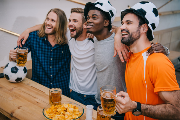 excited multicultural group of male football fans in soccer ball hats holding beer and watching soccer match at bar - Фото, зображення