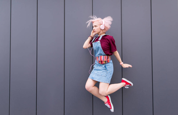 Girl with a funny haircut, headphones and stylish colored clothes jumping against the background of a dark wall. Outdoor portrait bright girl in a jump on a gray background - Photo, Image