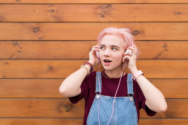 Счастливая, стильная девушка с розовыми волосами слушает музыку в розовых наушниках на фоне деревянной стены и смотрит в сторону. Смешная девочка-подросток на деревянном фоне
. - Фото, изображение