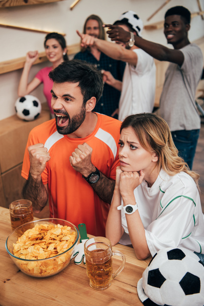 aficionados al fútbol multicultural emocional señalando con los dedos y viendo el partido de fútbol en el bar con patatas fritas y vasos de cerveza
  - Foto, Imagen