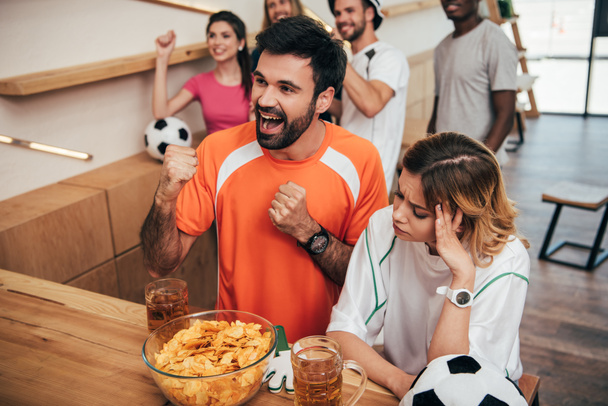 Lächelnder junger Mann in orangefarbenem Fan-T-Shirt feiert und macht Ja-Gesten, während seine aufgebrachte Freundin mit der Hand an der Bar mit Chips und Bier sitzt  - Foto, Bild
