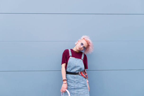 Stylish fashion girl with pink hair and denim clothing poses against the backdrop of a blue wall. Street portrait of a cute girl on a blue background - Foto, Imagem