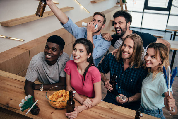 vista de ángulo alto de amigos multiculturales emocionados celebrando con cuernos de ventilador y palmadas de mano durante el reloj del partido de fútbol en el bar
  - Foto, imagen